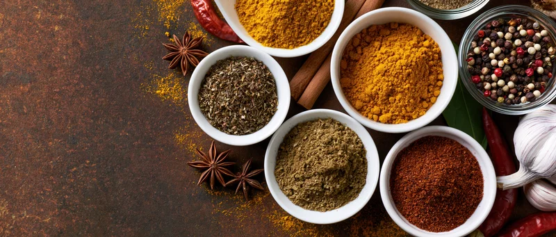 Assorted spices in white bowls and glass jars arranged on a dark rusted metal surface with copyspace on the left