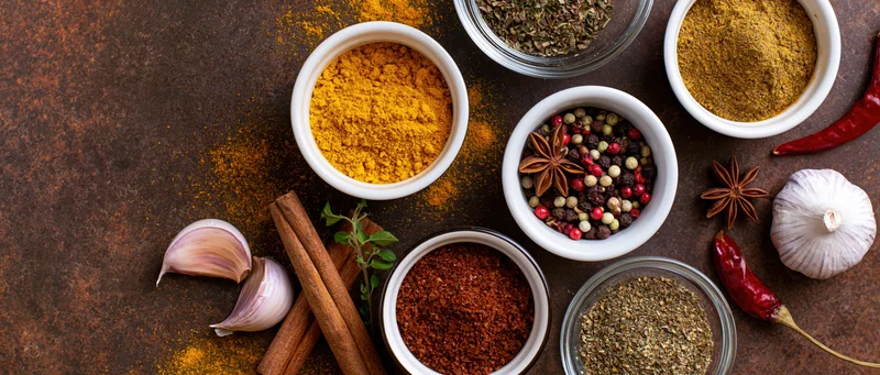 Assorted cooking spices in bowls and whole forms arranged on a dark rusted metal surface