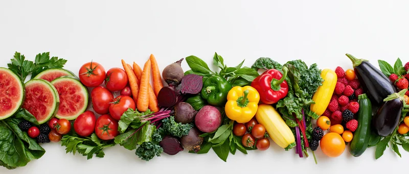 A row of assorted fresh fruits and vegetables arranged horizontally on a white background