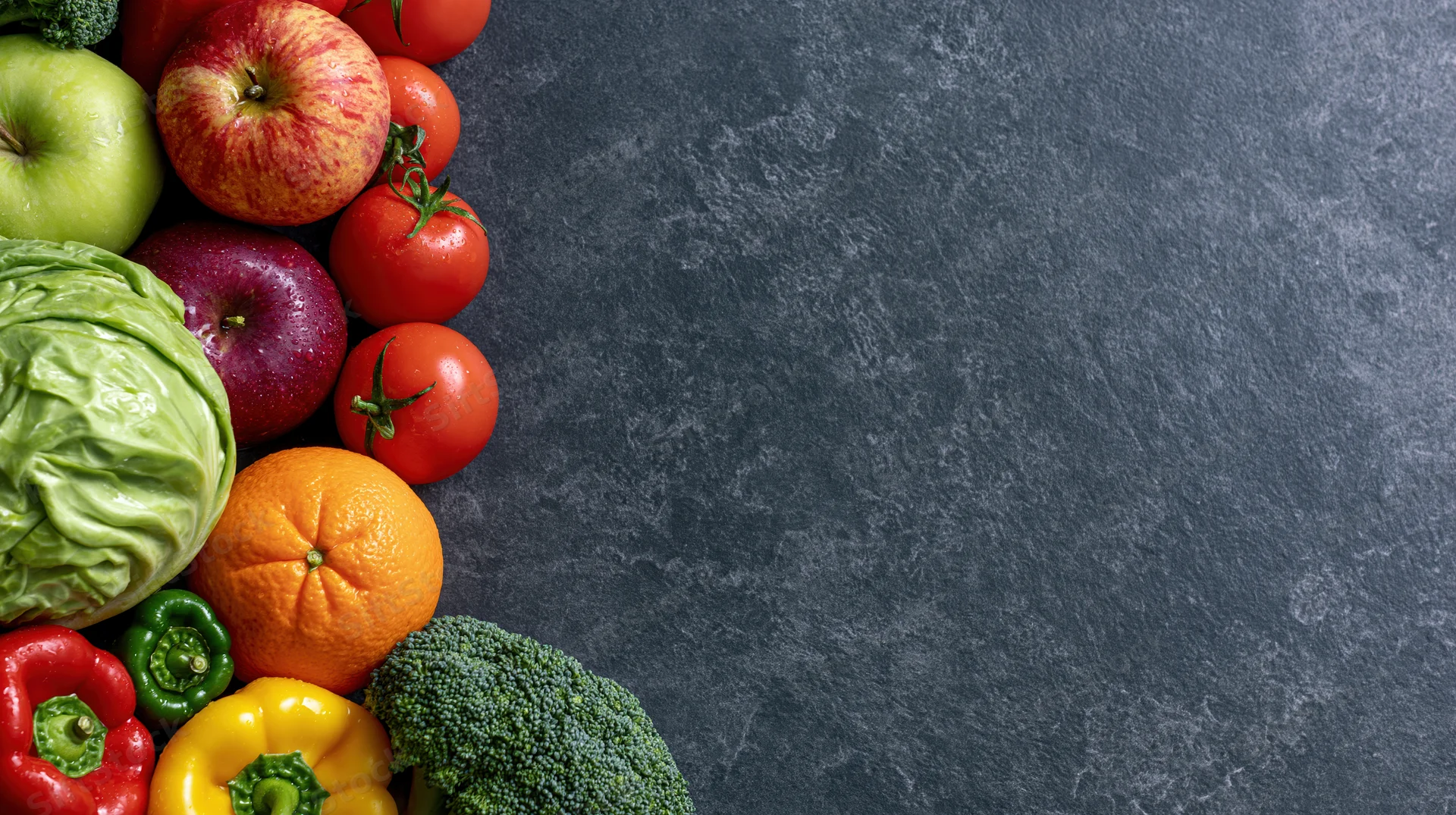 Fresh vegetables and fruits arranged along the left side of a dark slate surface
