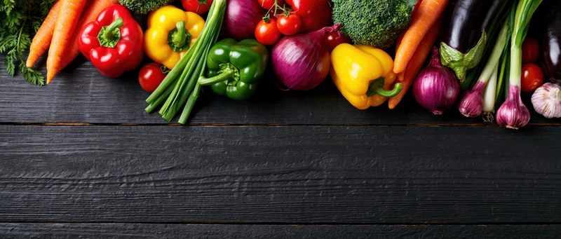Assorted fresh vegetables arranged along the top of a dark wooden surface with copyspace below
