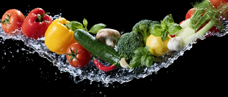 Fresh vegetables arranged in a curved line with water splashing around them on black background
