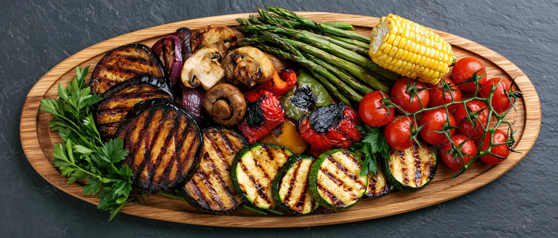 Assorted grilled vegetables arranged on an oval wooden serving platter on slate surface
