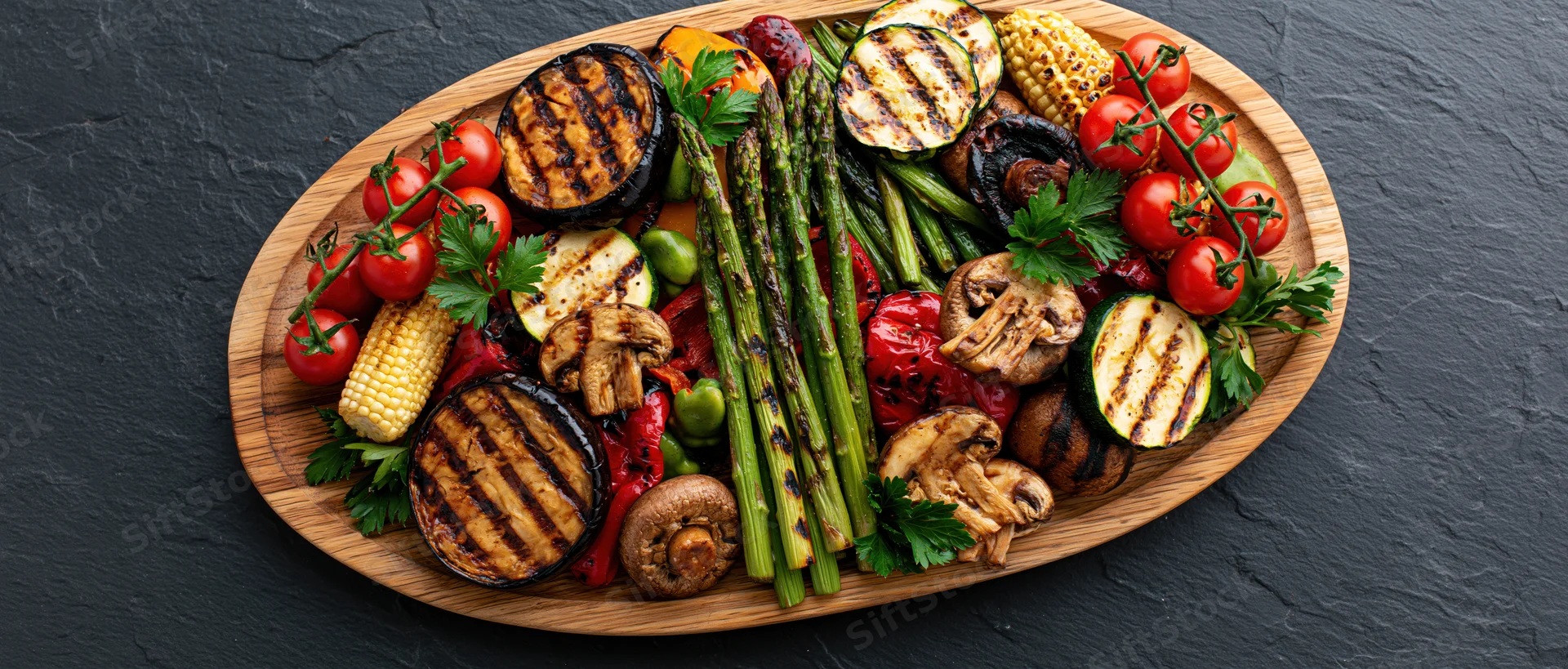 Assorted grilled vegetables arranged on an oval wooden serving platter on dark slate surface