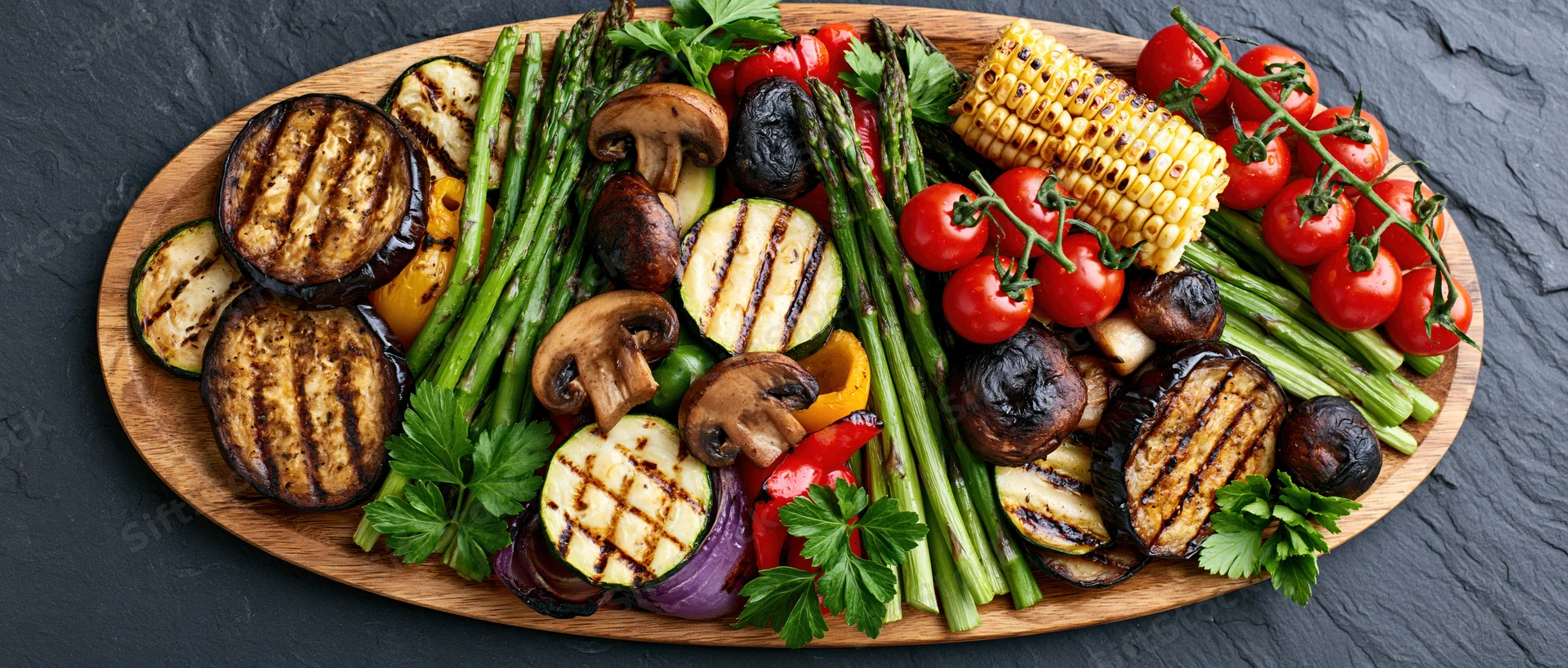 Grilled vegetables arranged on an oval wooden serving platter on a dark slate surface