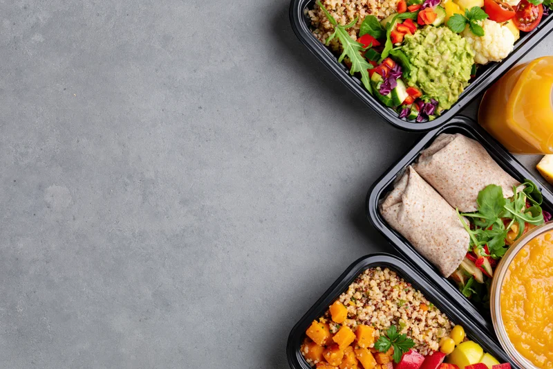 Healthy meal prep containers with vegetables, quinoa, wraps and juice arranged on gray countertop