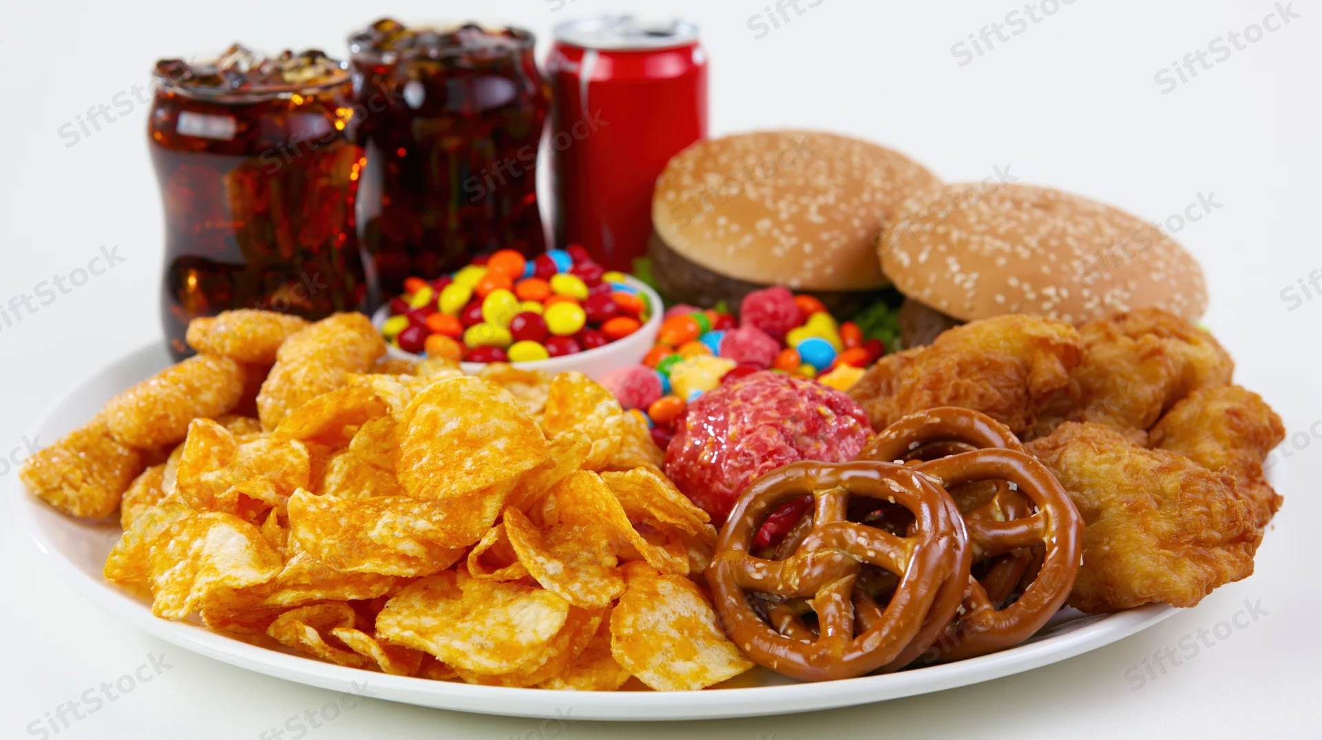 Assorted junk food including chips, burgers, soda, candy and fried chicken arranged on a white plate