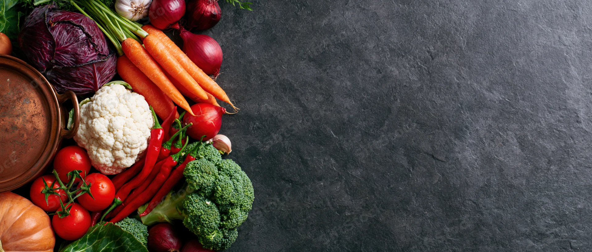 Assorted raw vegetables arranged along the left edge of a dark slate surface