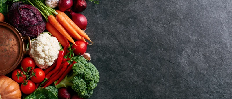 Assorted raw vegetables arranged along the left edge of a dark slate surface