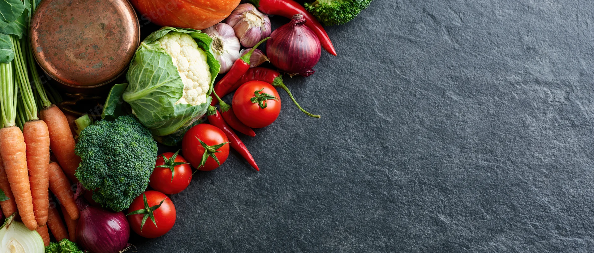 Assorted raw vegetables arranged along the left edge of a dark slate surface
