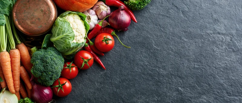 Assorted raw vegetables arranged along the left edge of a dark slate surface