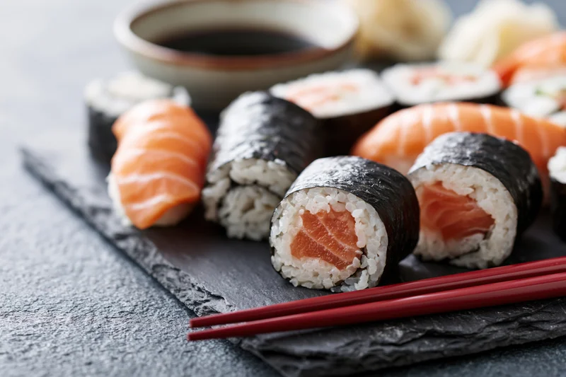 Assorted sushi rolls and nigiri pieces arranged on a dark slate plate with soy sauce and red chopsticks