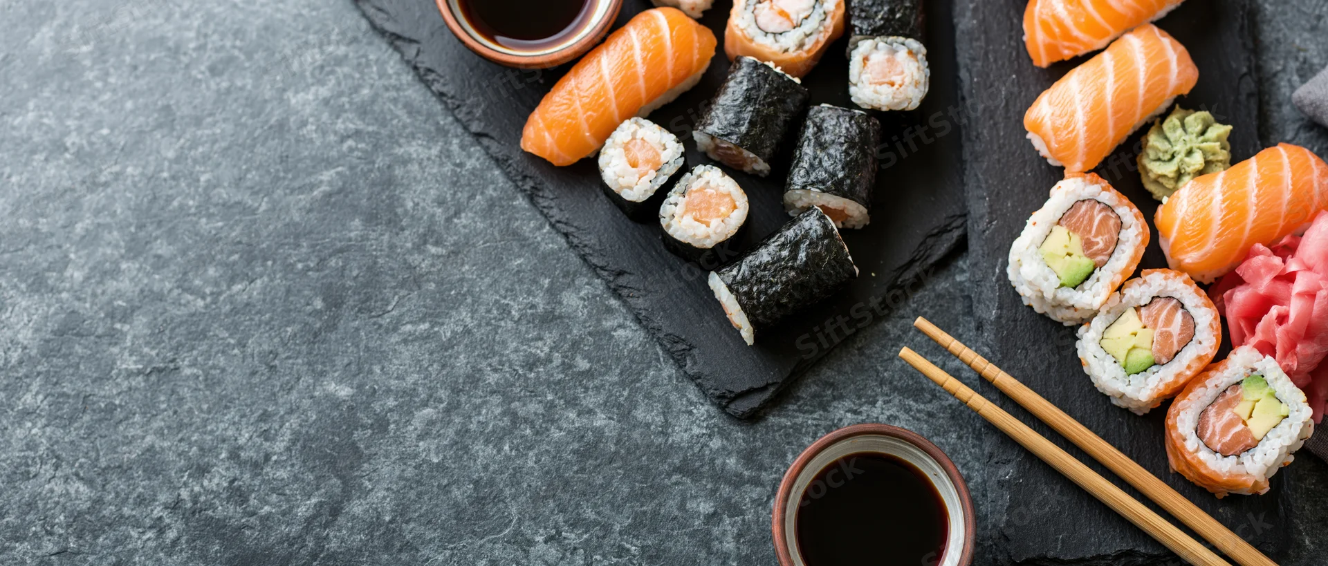 Assorted sushi with salmon and avocado rolls on black slate boards with soy sauce and chopsticks, overhead view