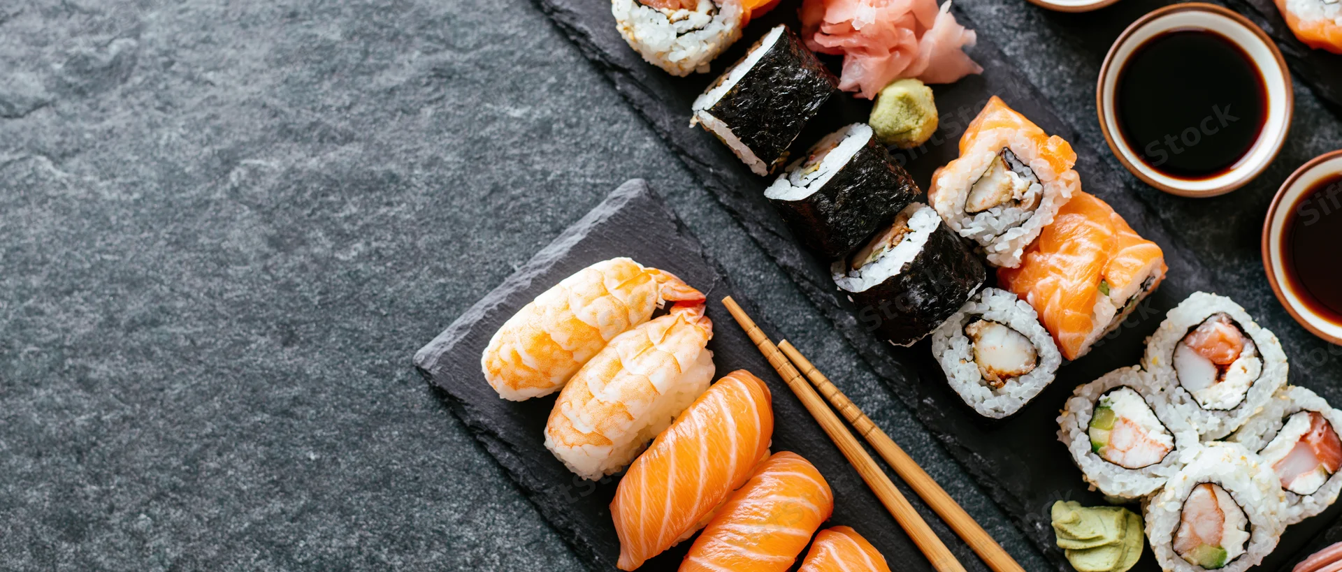Assorted sushi pieces with chopsticks and soy sauce arranged on black slate boards on a dark stone surface