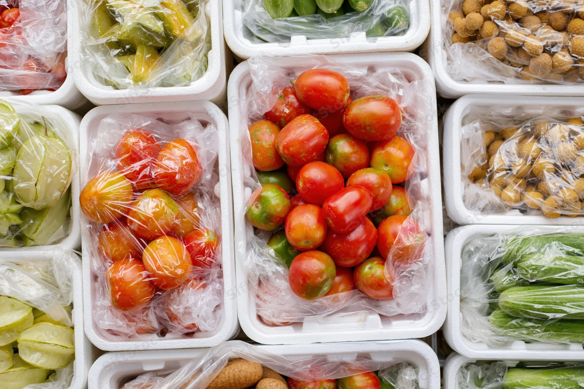 Assorted vegetables and fruits in plastic-wrapped foam containers on a market display
