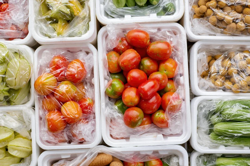 Assorted vegetables and fruits in plastic-wrapped foam containers on a market display