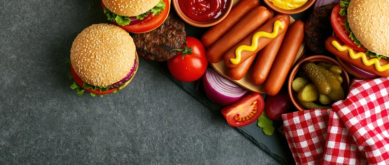 Barbecue cookout spread with burgers, hotdogs, and condiments arranged in corner on dark slate surface