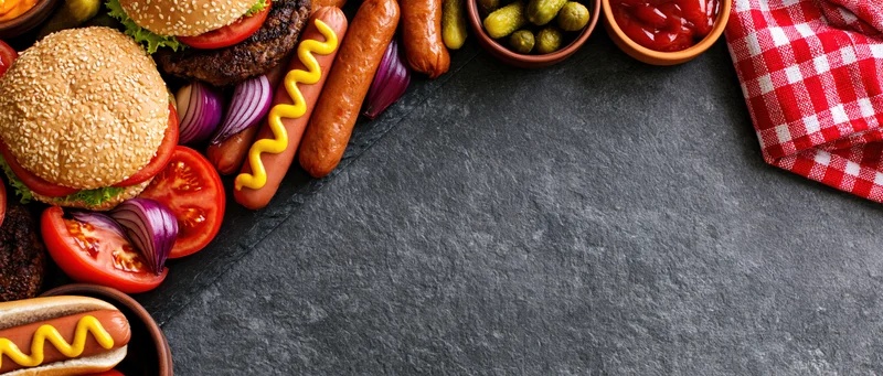 Barbecue cookout spread with hotdogs, hamburgers, sausages, condiments and checkered napkin arranged in left corner on dark slate surface