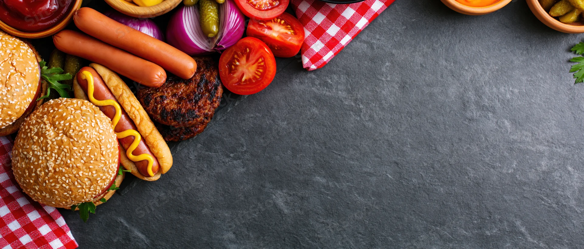 Barbecue cookout spread with grilled hotdogs, hamburgers, vegetables and condiments arranged in the left corner on dark slate surface with copyspace