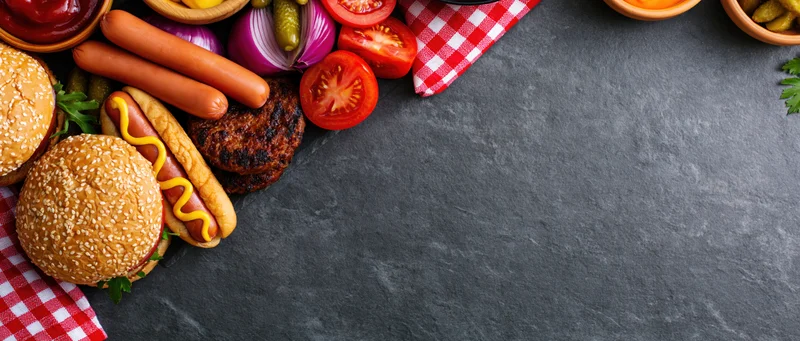 Barbecue cookout spread with grilled hotdogs, hamburgers, vegetables and condiments arranged in the left corner on dark slate surface with copyspace
