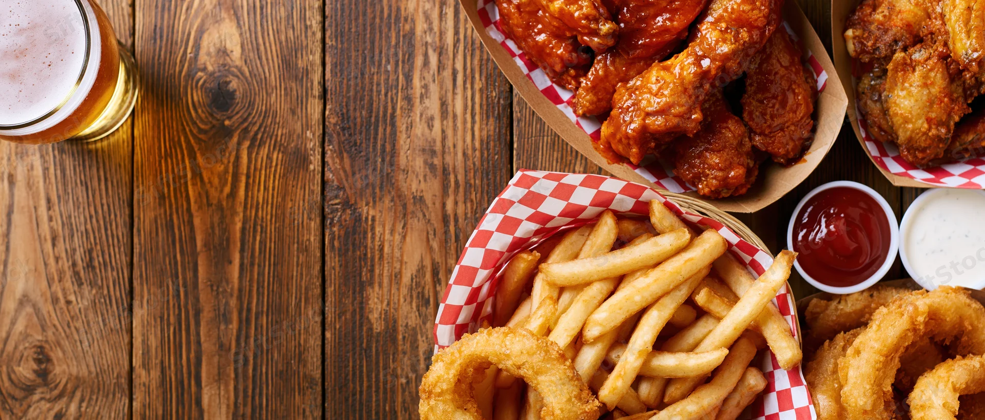Bar food spread with buffalo chicken wings, fries, onion rings, and beer on a worn wooden plank table