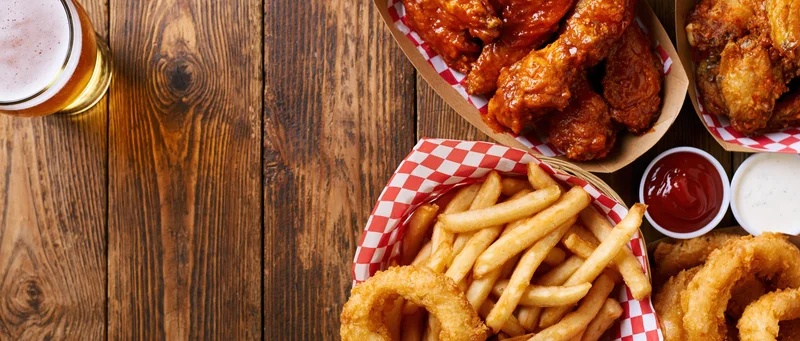 Bar food spread with buffalo chicken wings, fries, onion rings, and beer on a worn wooden plank table