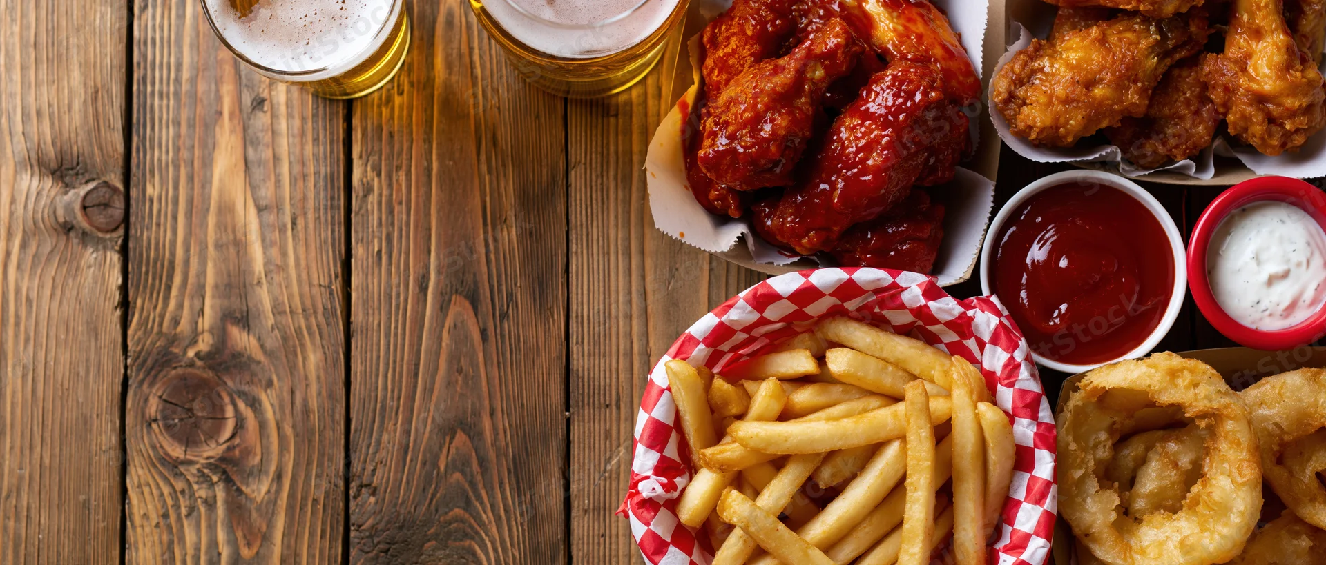 Bar food spread with buffalo chicken wings, fries, onion rings, and beer on a worn wooden plank table