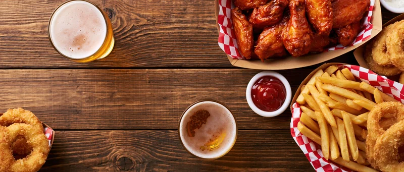 Buffalo chicken wings, fries, onion rings and beer arranged on a worn wooden plank table with ketchup and sauce in small bowls