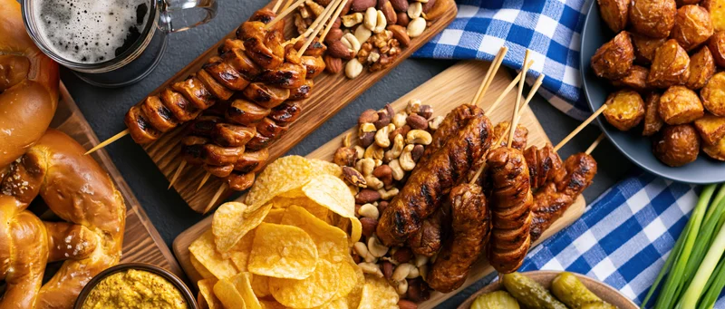 Bavarian Oktoberfest food spread with sausages, pretzels, chips, nuts and beer arranged on wooden boards and a checkered cloth over a dark grey surface