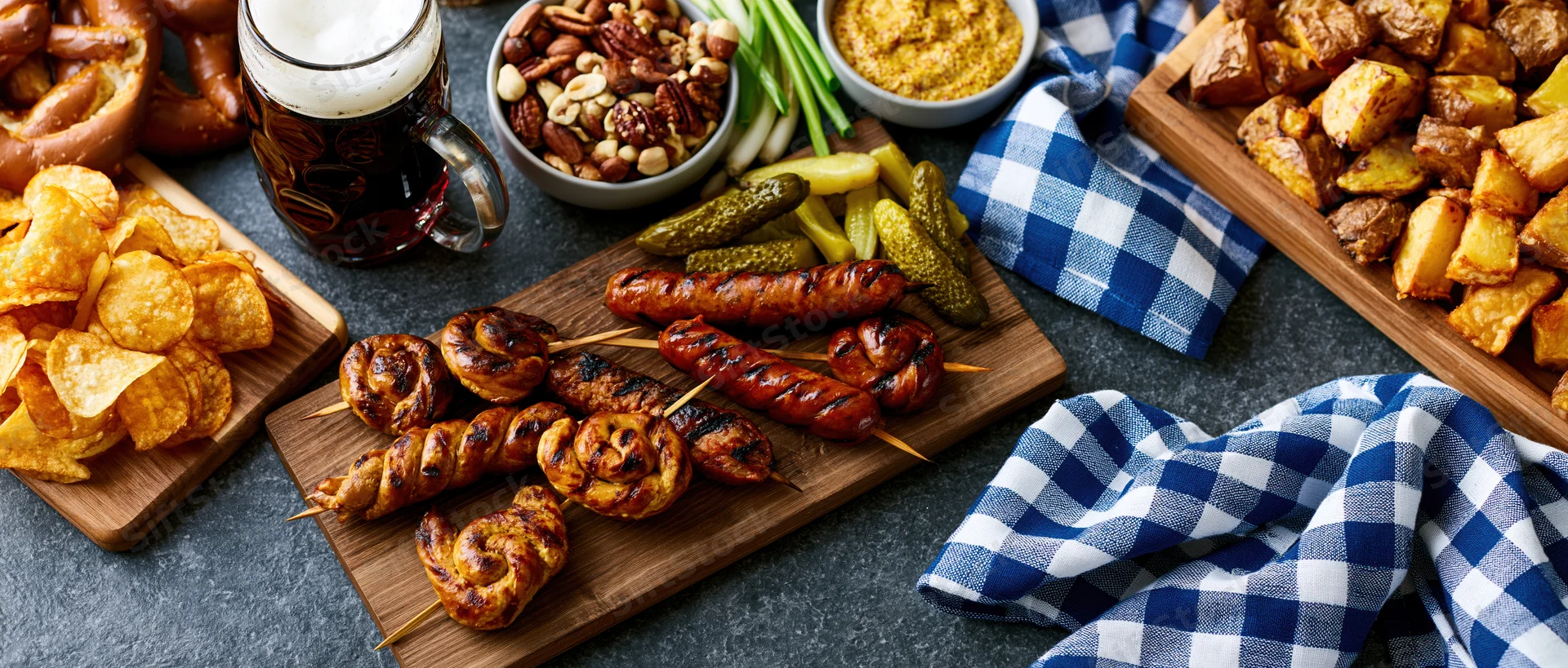 Bavarian food spread with grilled sausages, pretzels, chips, beer and side dishes arranged on dark table with checkered napkins