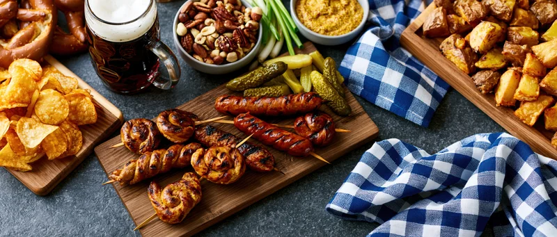 Bavarian food spread with grilled sausages, pretzels, chips, beer and side dishes arranged on dark table with checkered napkins