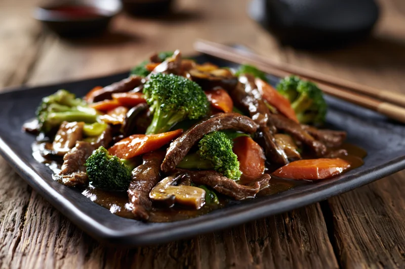 Beef and vegetable stir-fry served on a dark square ceramic plate with chopsticks on a wooden table