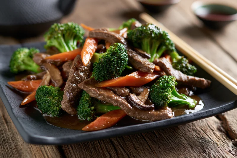 Beef and vegetable stir-fry served on a dark square ceramic plate with chopsticks on a wooden table