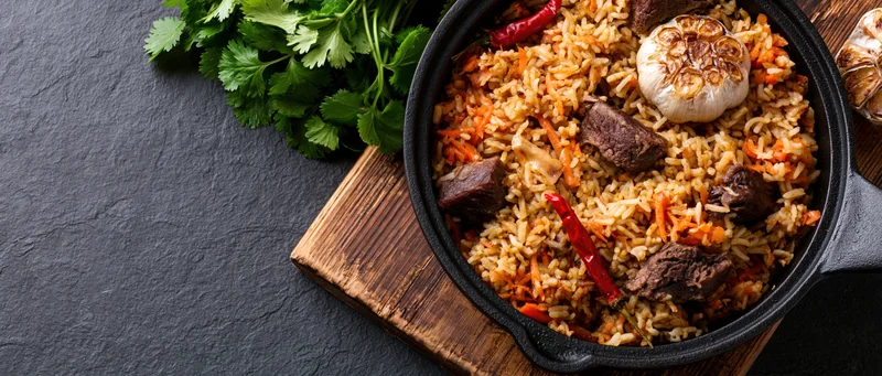 Central asian plov rice pilaf with beef and vegetables in a cast iron pan on a wooden board