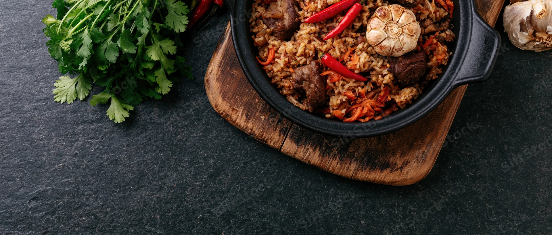 Central asian plov rice pilaf in a black cast iron pan on a wooden board with fresh herbs and vegetables nearby