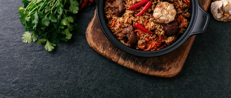 Central asian plov rice pilaf in a black cast iron pan on a wooden board with fresh herbs and vegetables nearby