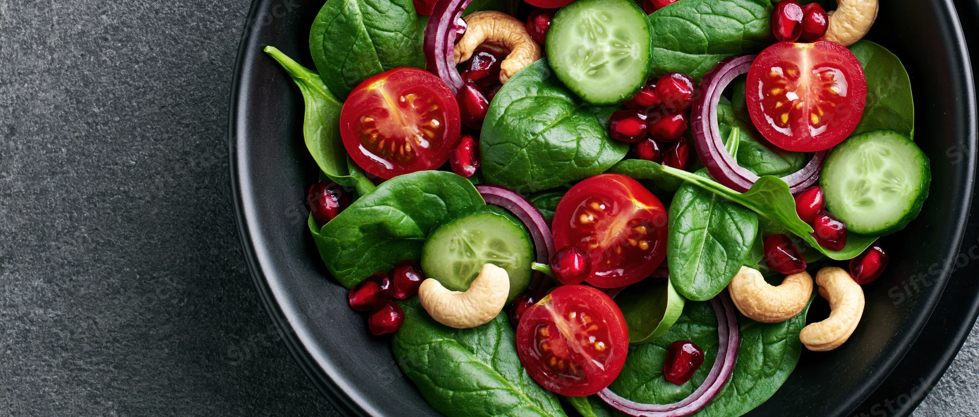 Fresh spinach salad with tomatoes cucumbers pomegranate and cashews in a black ceramic bowl on dark surface