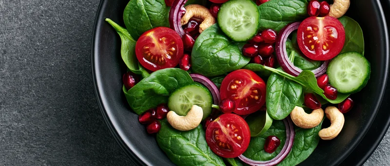 Fresh spinach salad with tomatoes cucumbers pomegranate and cashews in a black ceramic bowl on dark surface