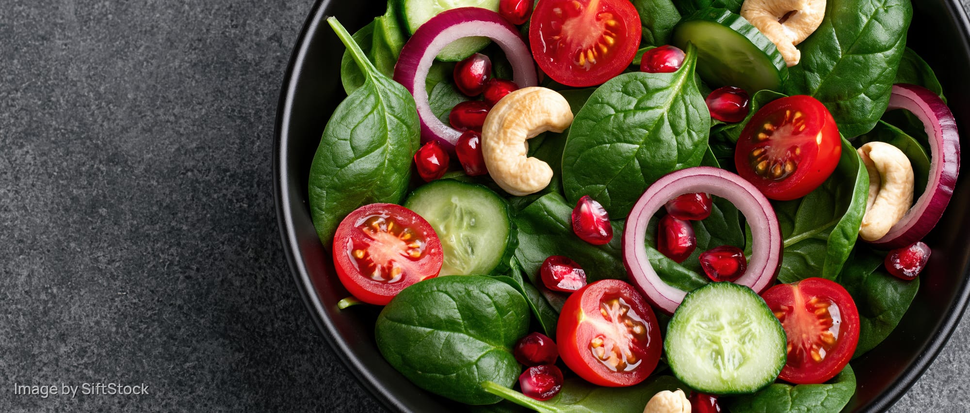 Fresh spinach salad with tomatoes cucumbers onions pomegranate and cashews in a black ceramic bowl on dark surface