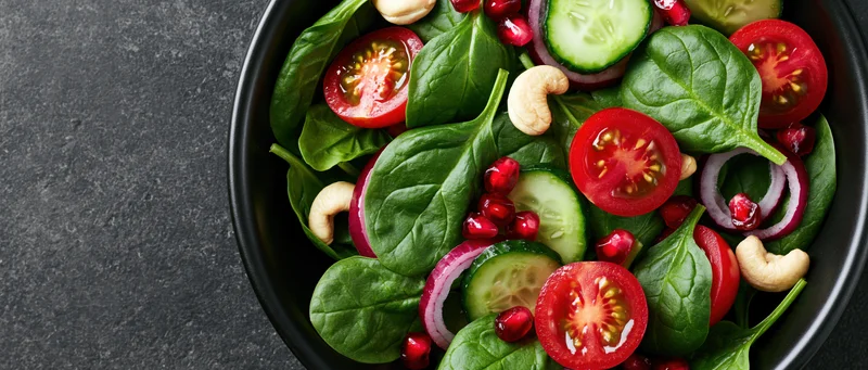 Fresh spinach salad with tomatoes cucumbers onions pomegranate and cashews in black ceramic bowl on dark surface