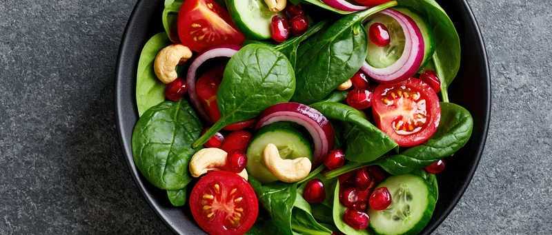 Fresh spinach salad with tomatoes cucumbers onions pomegranate and cashews in a black ceramic bowl on dark surface