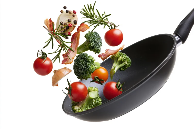 Black non-stick frying pan tilted mid-toss with flying vegetables and bacon against white background