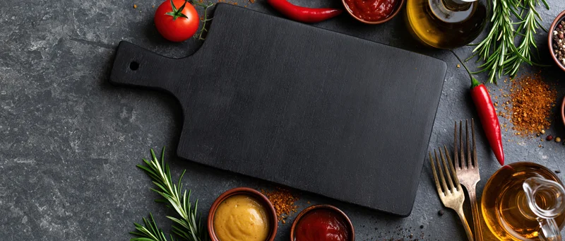 Black wooden cutting board surrounded by fresh ingredients and kitchen tools on dark stone surface