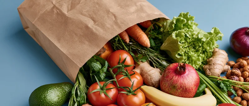 Brown paper grocery bag tipped on its side spilling fresh produce and vegetables onto a blue surface