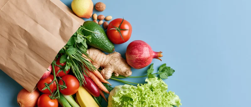 Brown paper grocery bag tipped over spilling fresh vegetables and fruits on blue surface