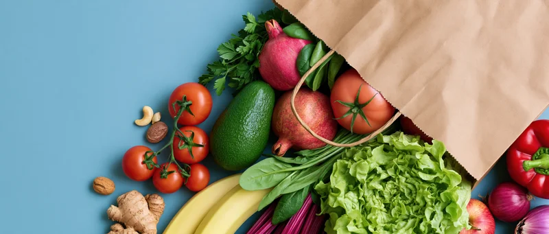 Brown paper grocery bag tipped over spilling fresh vegetables and fruits on blue surface
