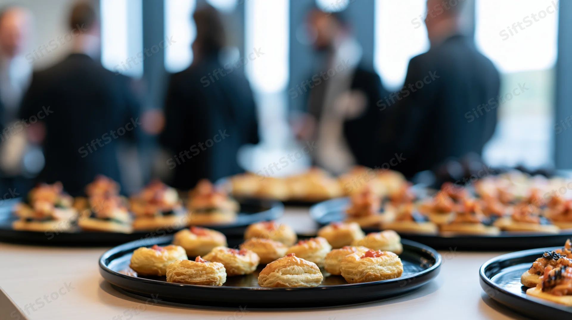 Small appetizers arranged on black trays on a table with blurred people in business attire in the background