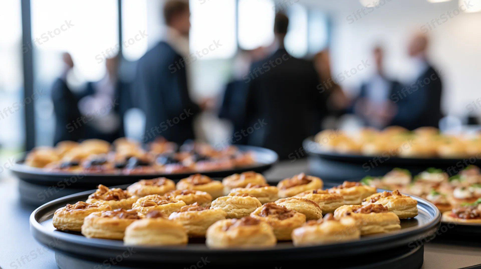 Golden puff pastry canapes on black trays at a corporate event buffet with professionals in suits in the background