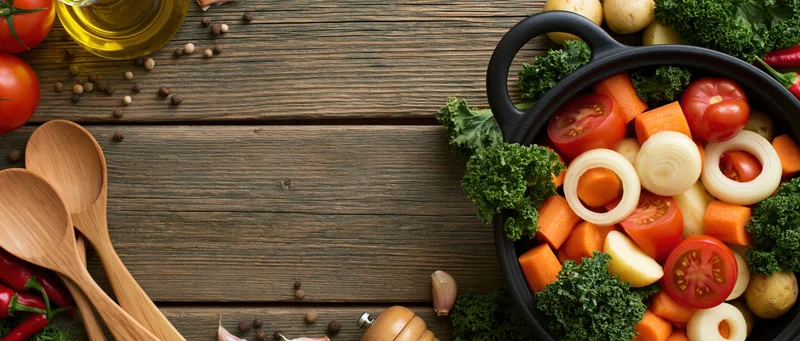 Cast iron pot filled with chopped raw vegetables on wooden table with cooking utensils and ingredients around