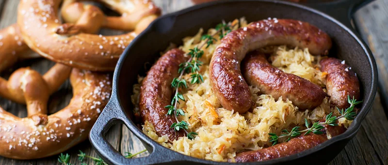 Bratwurst sausages with sauerkraut in a cast iron skillet beside pretzels on a wooden table